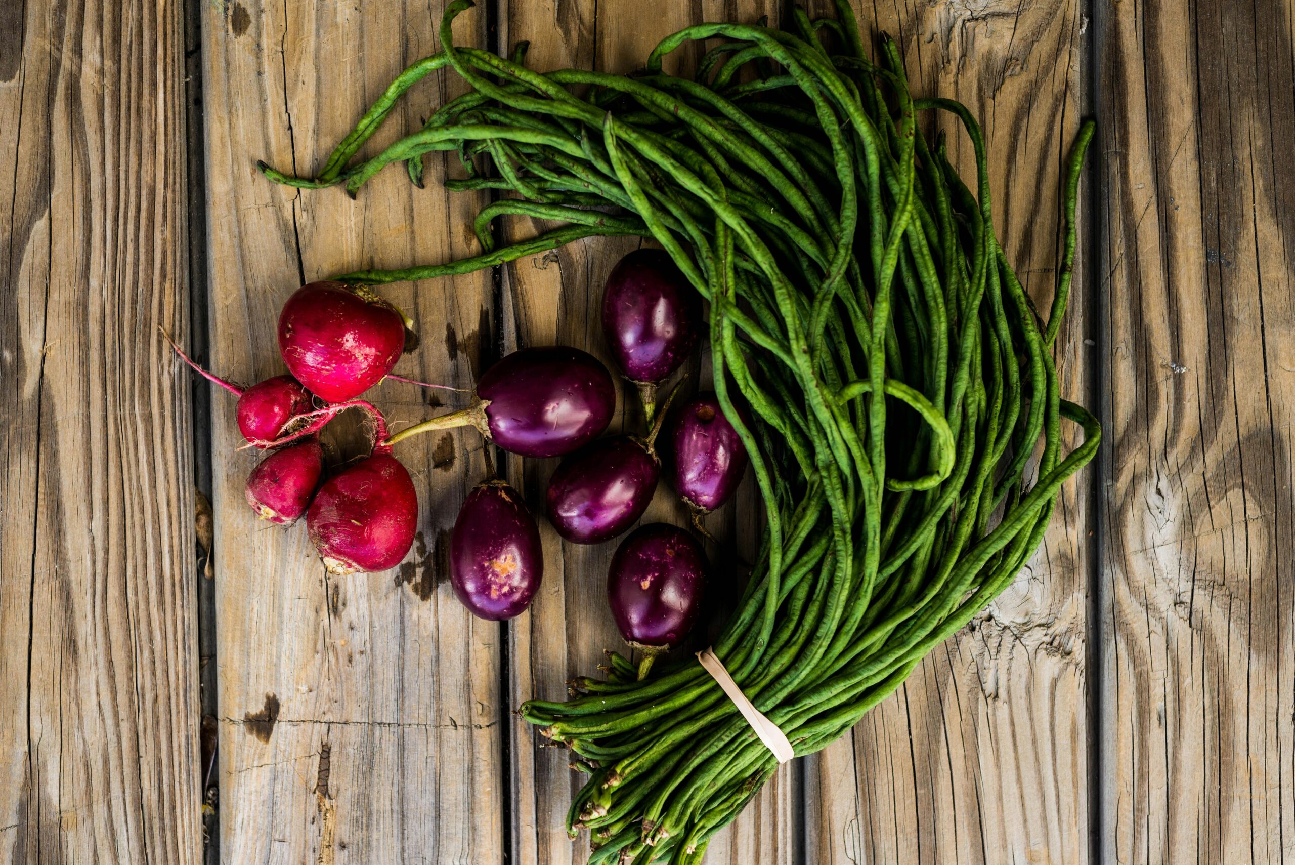 Top view of fresh radishes, eggplant, and yardlong beans on wooden table.